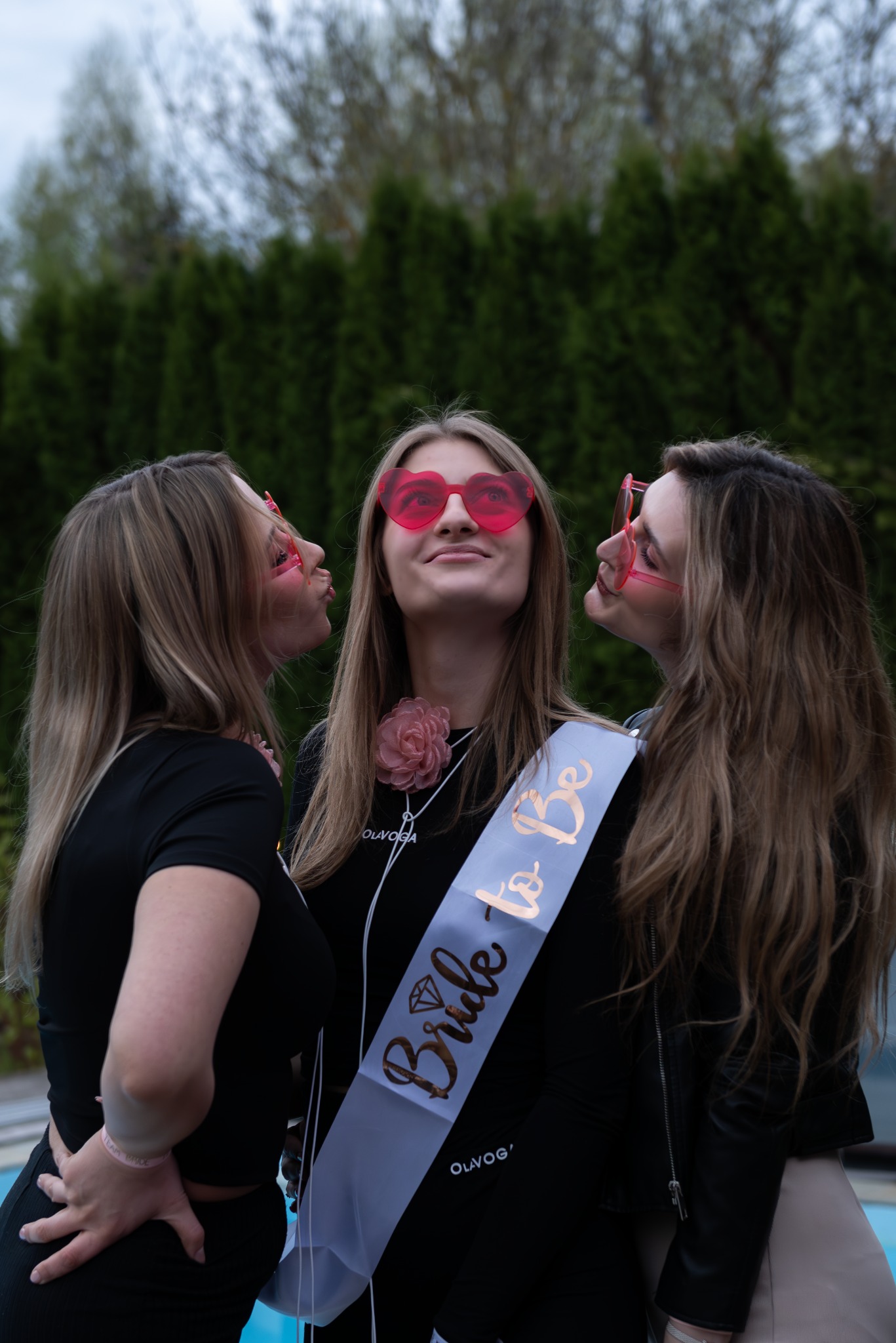 Trzy uśmiechnięte kobiety w okularach w kształcie serc, świętujące wieczór panieński, z szarfą 'Bride to Be' na tle zielonych drzew.