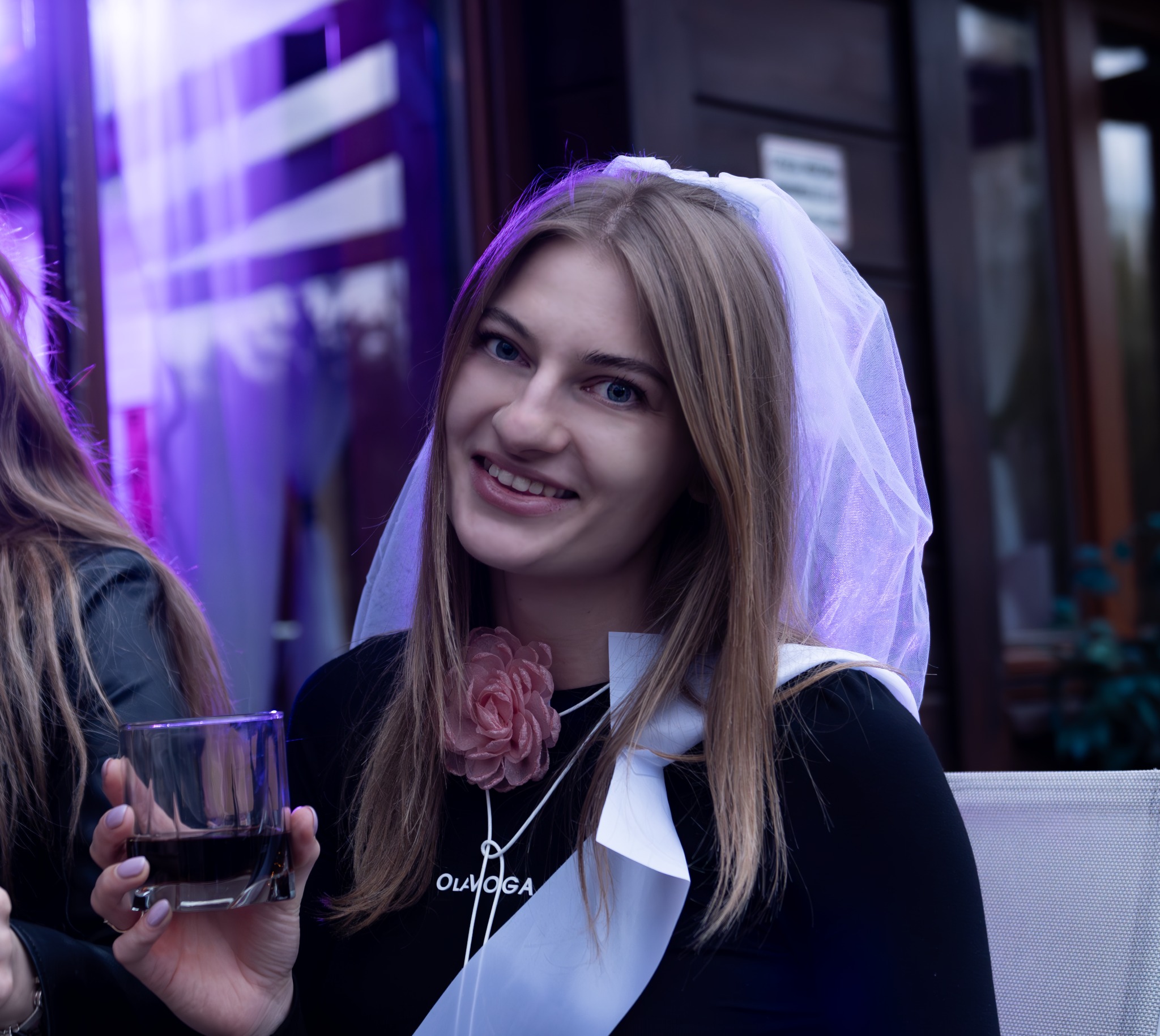 Uśmiechnięta panna młoda w welonie, z różową ozdobą i szarfą, trzyma drinka w szklance na tle fioletowego oświetlenia.