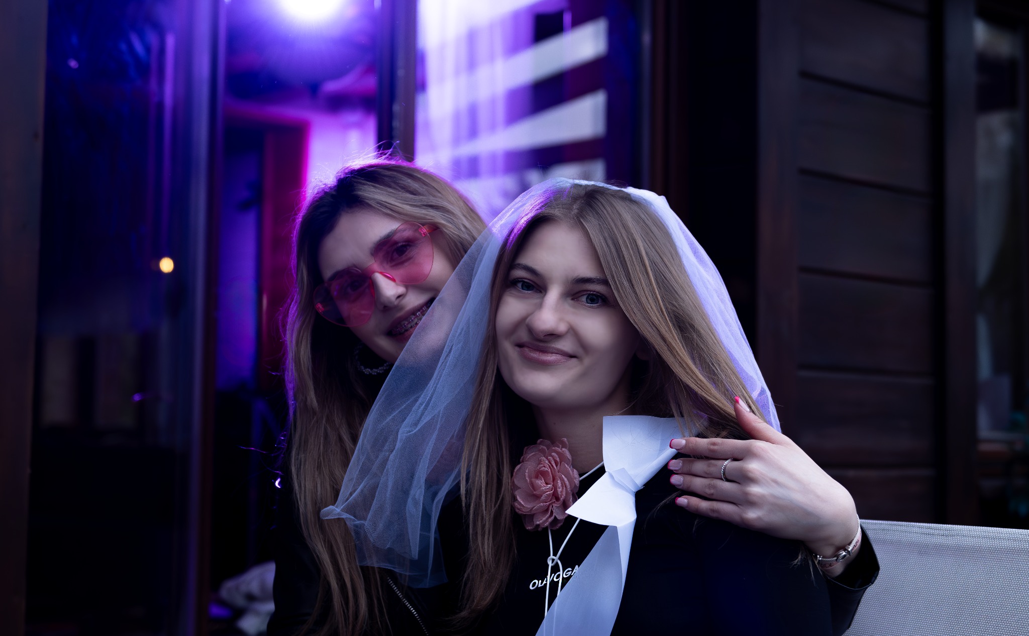 Uśmiechnięta panna młoda z welonem i ozdobnym kwiatem na szyi, obejmowana przez przyjaciółkę w różowych okularach w kształcie serc, pozuje na tle ciemnego, drewnianego budynku z oświetleniem...