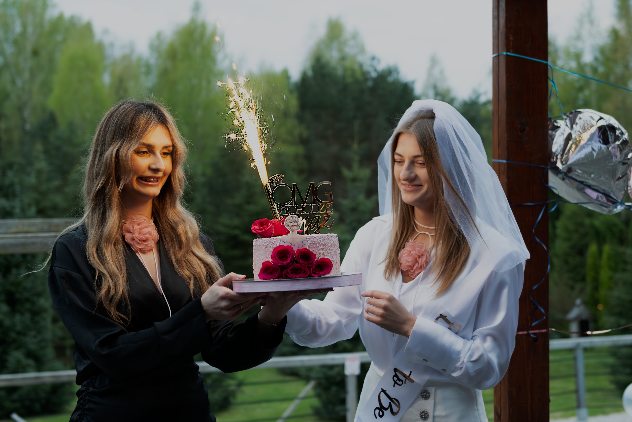 Dwie kobiety trzymają tort z fontanną iskier, jedna w czarnej sukience z broszką w kształcie kwiatu, druga w białej koszuli i welonie z szarfą 'Bride to Be'.