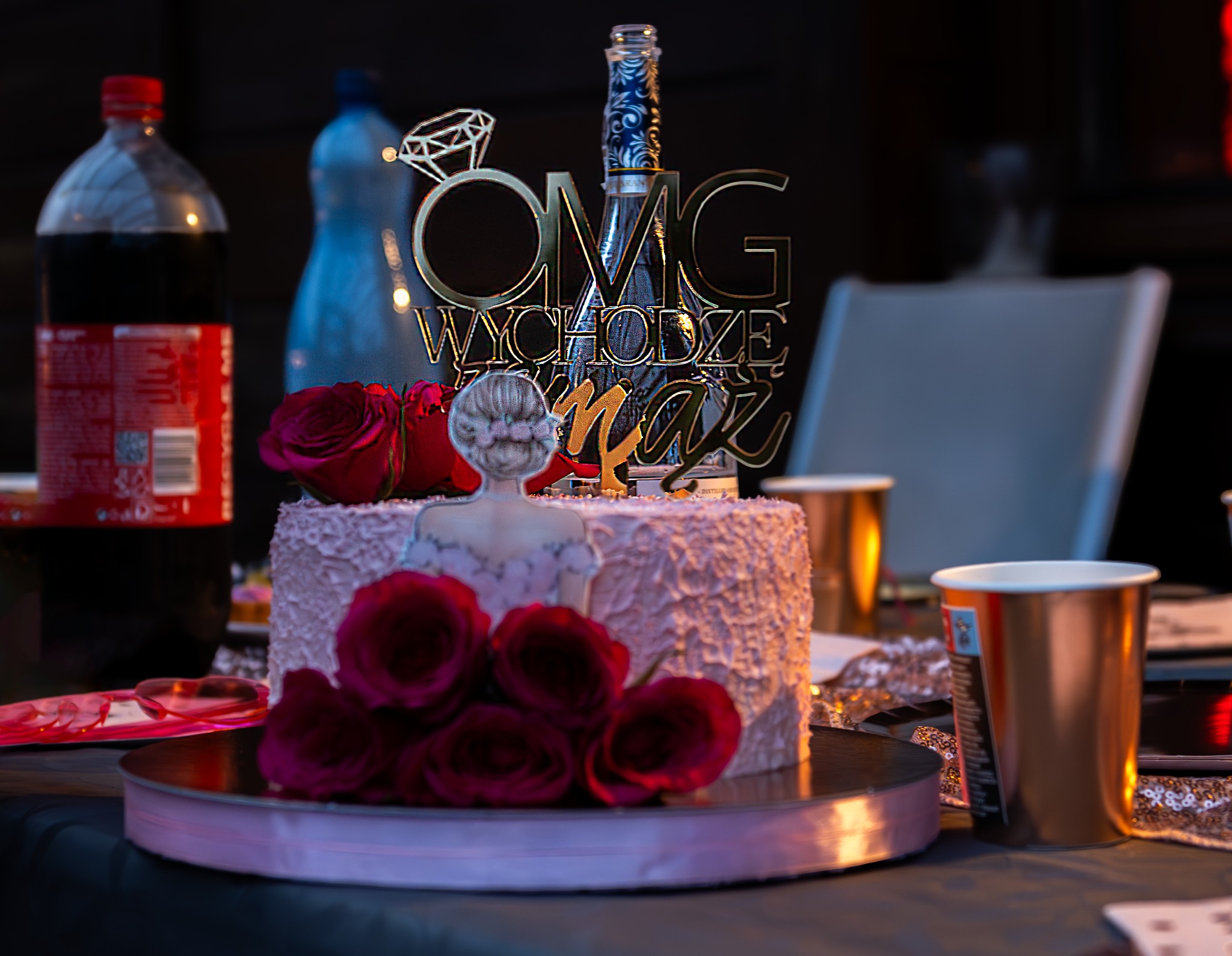 Tort z różowym kremem, ozdobiony czerwonymi różami i topperem 'OMG Wychodzę za mąż', na stole z dekoracjami.