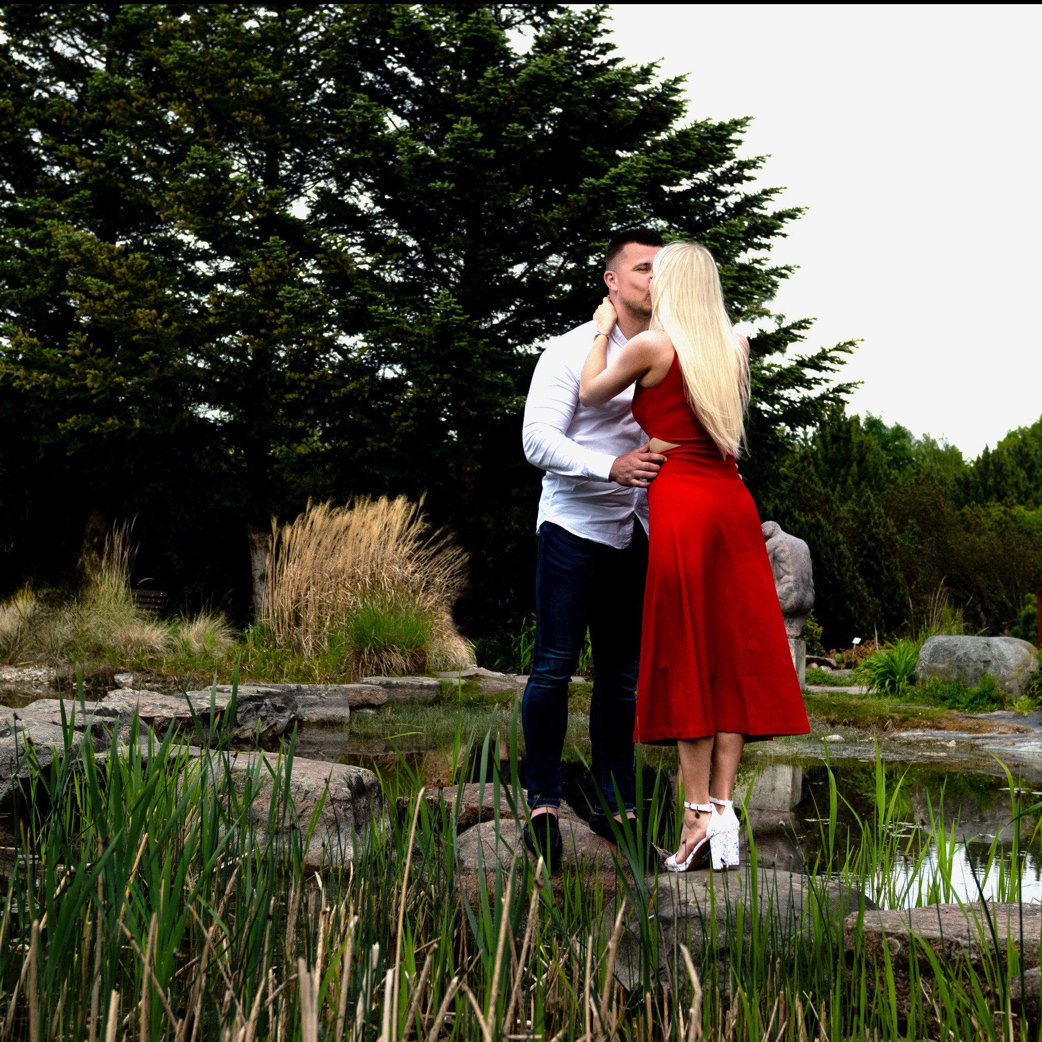 Para całująca się na tle parkowego stawu, kobieta w czerwonej sukience i białych butach na obcasie, mężczyzna w białej koszuli i dżinsach.