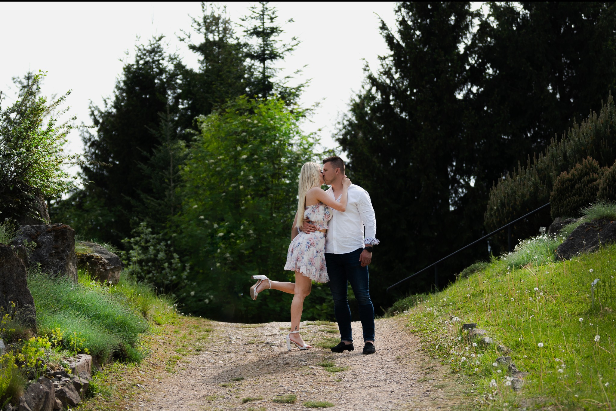 Para całująca się na leśnej ścieżce, kobieta w kwiecistej sukience i szpilkach, mężczyzna w białej koszuli i dżinsach, tło z drzew i zieleni.