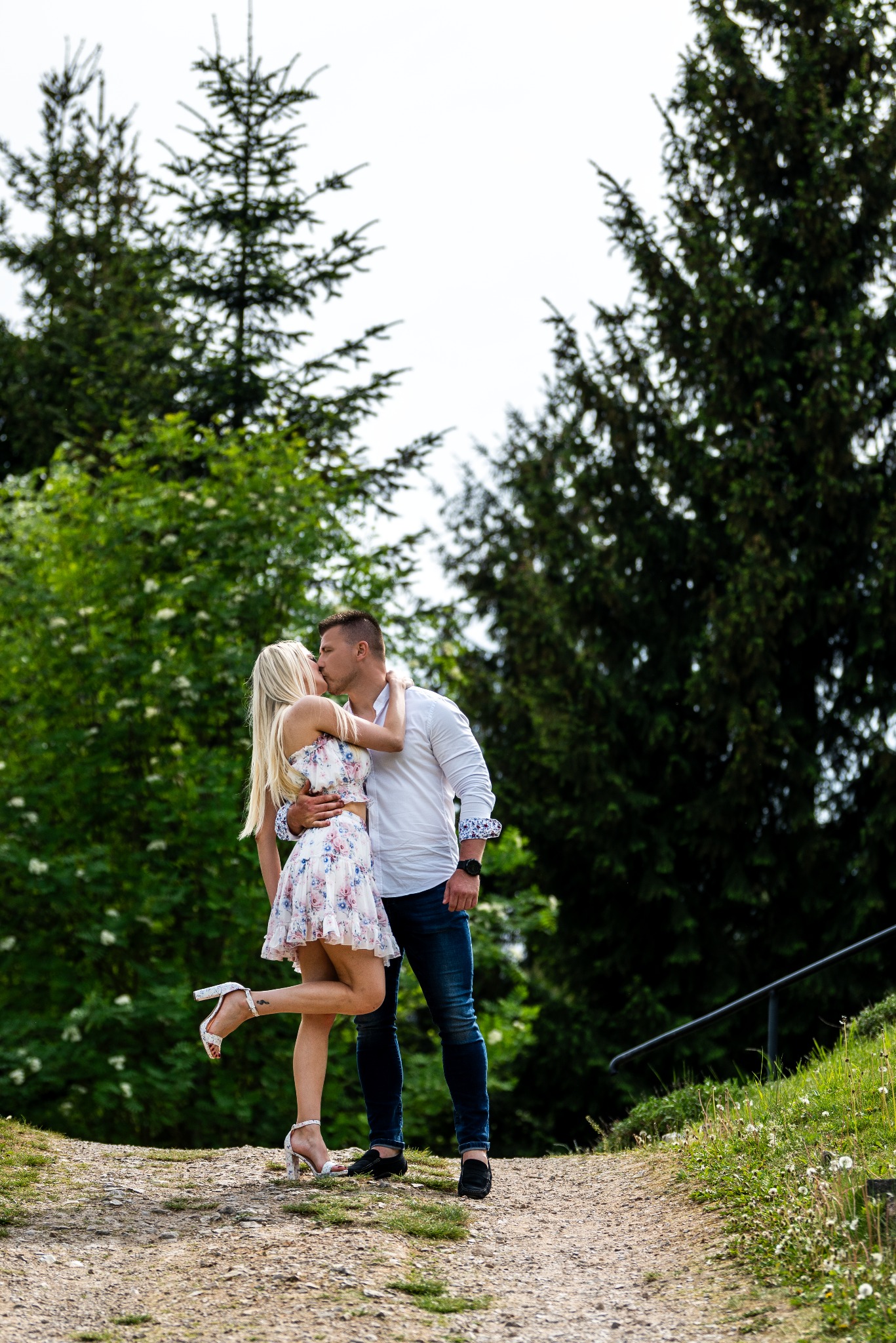 Para całuje się na leśnej ścieżce; kobieta w kwiecistej sukience i sandałach, mężczyzna w białej koszuli i jeansach, wysokie drzewa w tle.