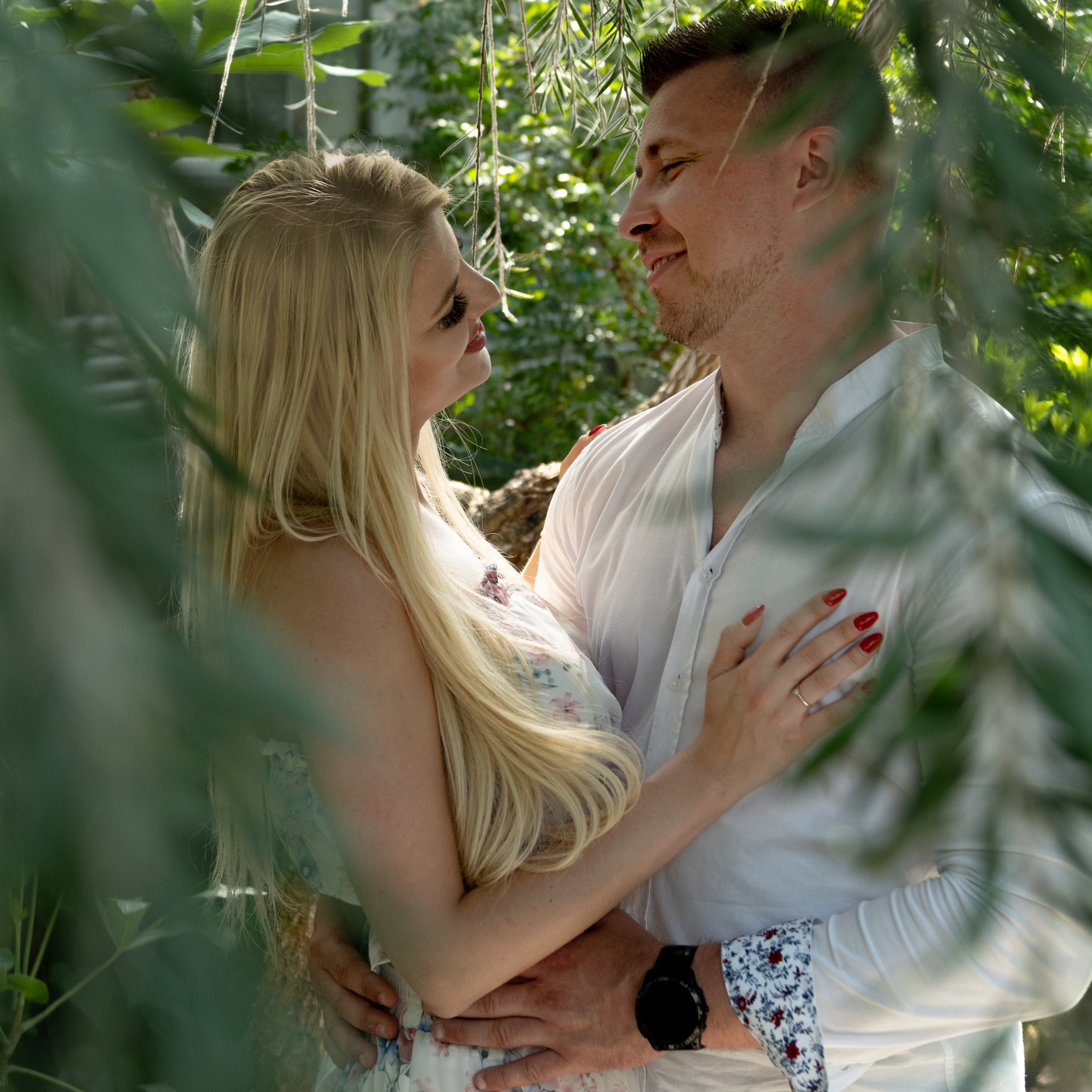 Para zakochanych, kobieta w kwiecistej sukience i mężczyzna w białej koszuli, obejmują się na tle zieleni, patrząc sobie w oczy.