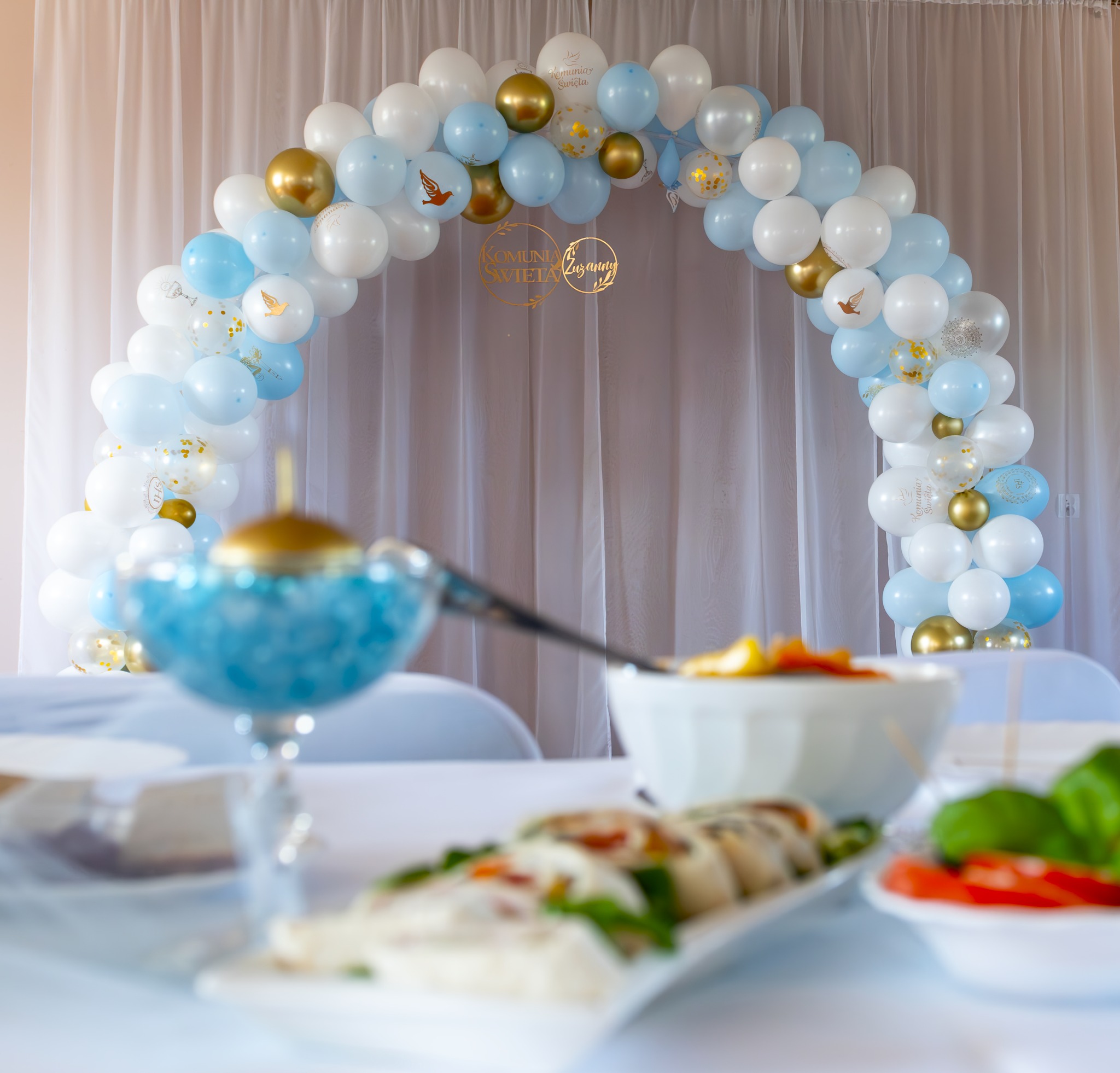 Dekoracja balonowa w odcieniach bieli i błękitu z elementami złota, tworząca łuk nad stołem z przekąskami, przygotowana na uroczystość Komunii Świętej.