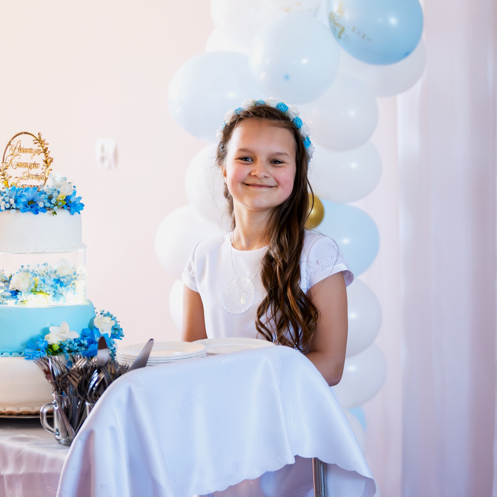 Uśmiechnięta dziewczynka w białej sukience i wianku z niebieskimi kwiatami, pozuje obok tortu komunijnego z niebieskimi dekoracjami i balonów w odcieniach bieli i błękitu.