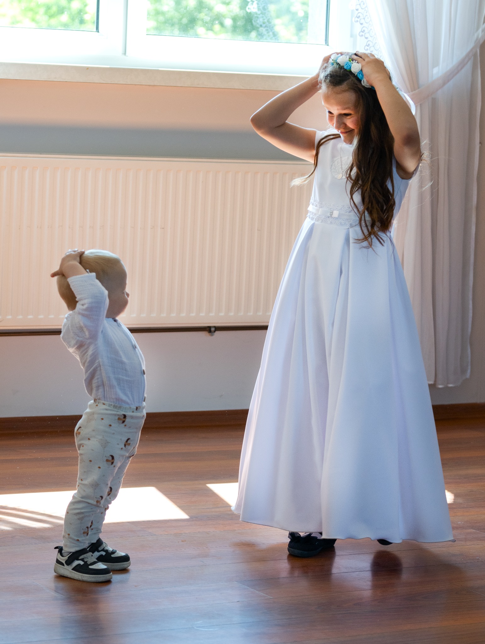 Dziewczynka w białej sukience i wianku na głowie, poprawia go, a mały chłopiec w koszuli i spodniach z nadrukiem, naśladuje ją, stojąc na drewnianej podłodze w jasnym wnętrzu.