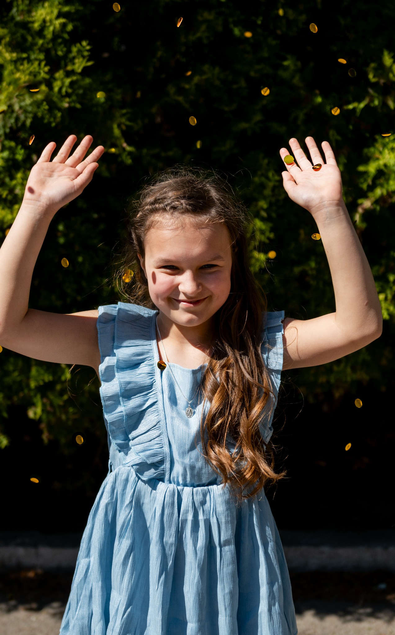 Uśmiechnięta dziewczynka w błękitnej sukience z falbankami, stojąca na tle zielonych krzewów i rzucająca w górę złote konfetti.