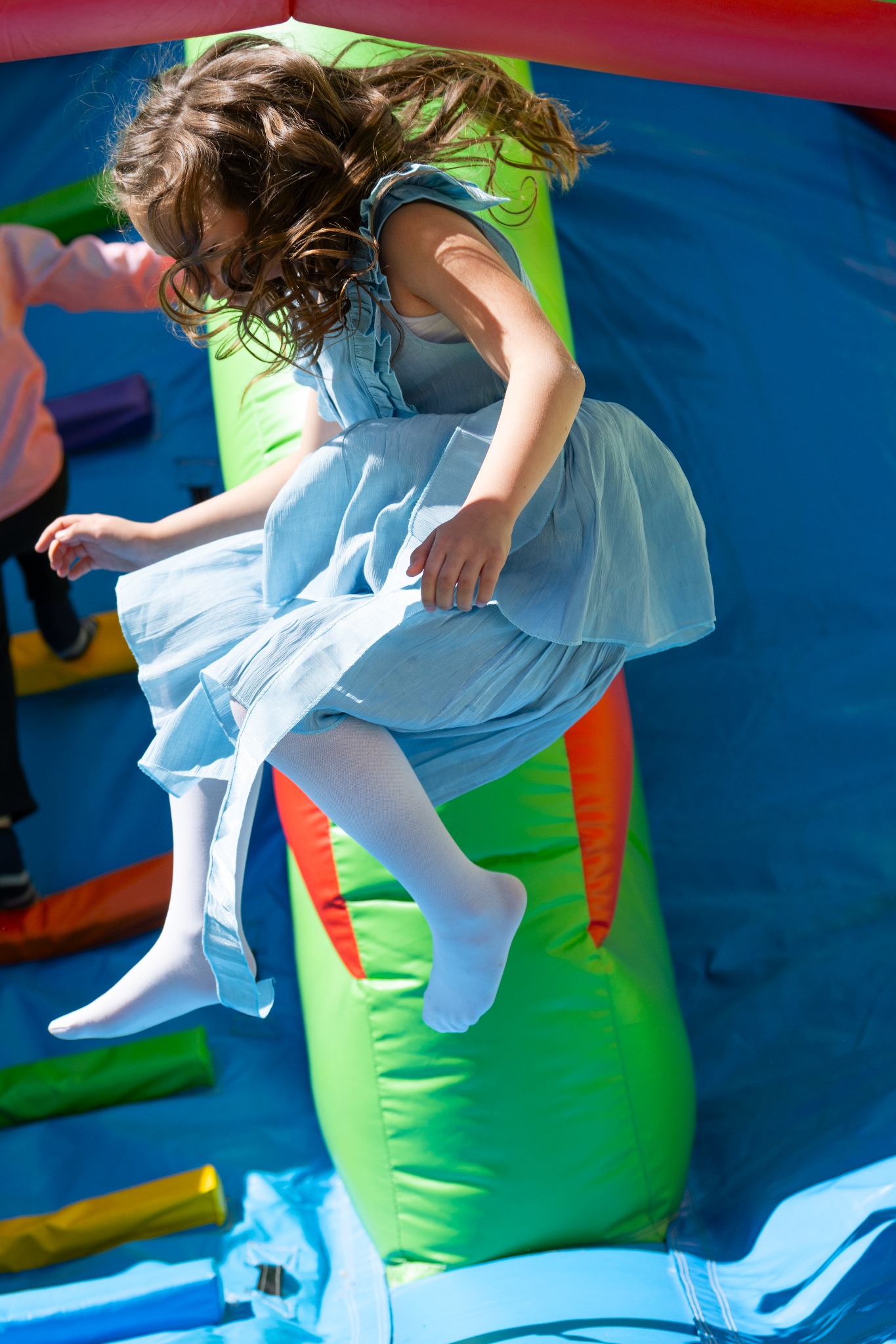 Dziewczynka w błękitnej sukience i białych rajstopach skacze na dmuchanym zamku, włosy rozwiane w powietrzu.