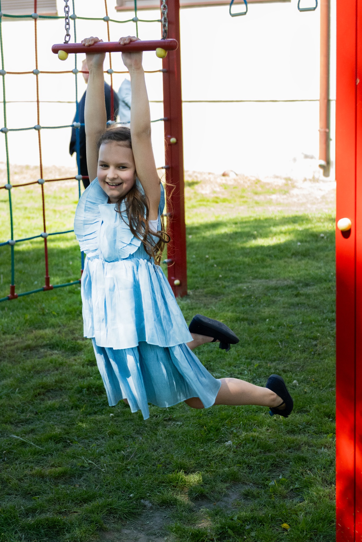 Uśmiechnięta dziewczynka w błękitnej sukience zwisa na rękach z drabinki na placu zabaw, z zieloną trawą w tle.