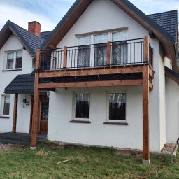 Wood System Adam Gajewski - Biały dom z drewnianym balkonem wspartym na drewnianych słupach, ozdobionym drobnymi lampkami, z czarną balustradą i numerem domu 22.