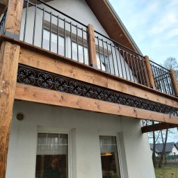 Wood System Adam Gajewski - Drewniany balkon z czarną, kutą balustradą, wsparty na solidnych, impregnowanych belkach, widoczny z perspektywy dolnej, z fragmentem elewacji budynku w tle.