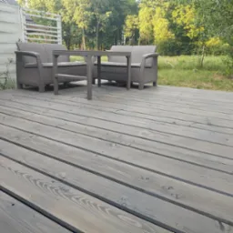 Szary taras z desek z widocznymi śrubami, na którym stoi zestaw mebli ogrodowych z technorattanu w odcieniach szarości, a w tle widać zieleń drzew i biały płot.