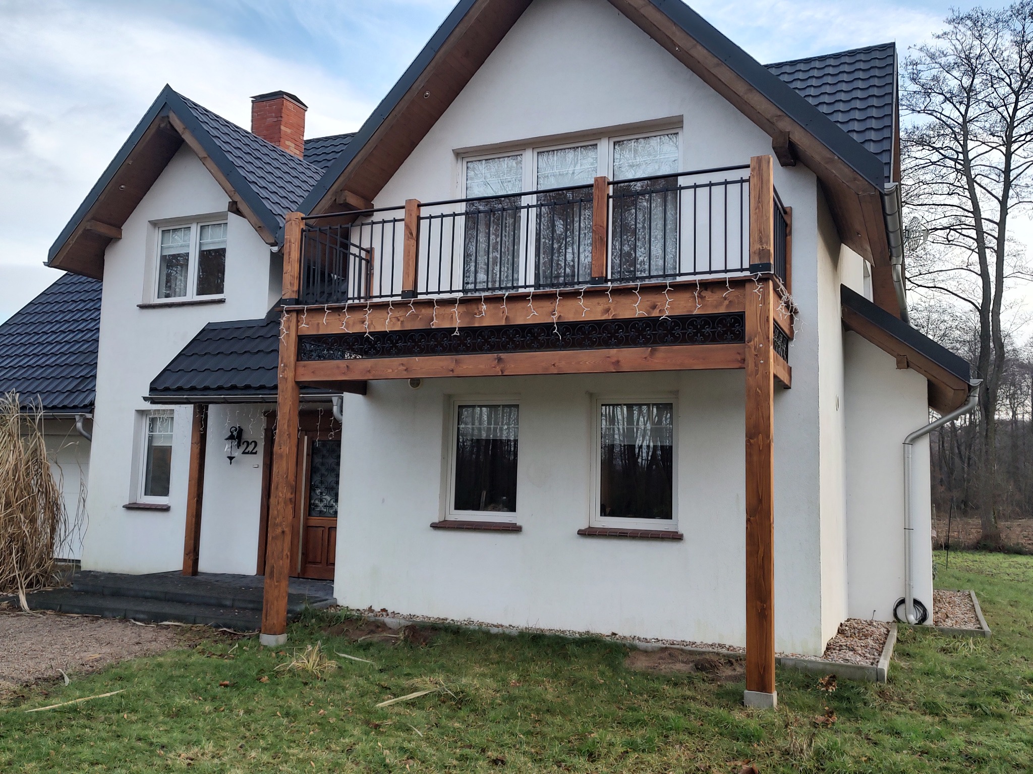 Biały dom z drewnianym balkonem wspartym na drewnianych słupach, ozdobionym drobnymi lampkami, z czarną balustradą i numerem domu 22.