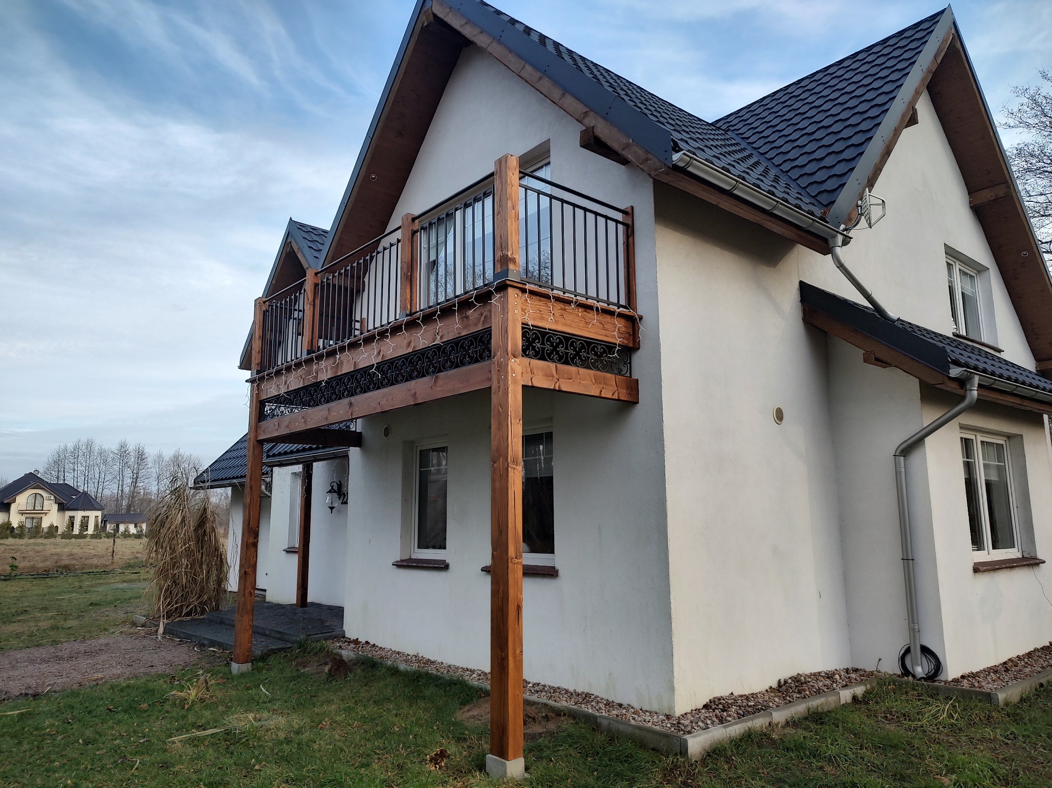 Biały dom z drewnianym balkonem z czarną, metalową balustradą, podparty drewnianymi słupami, z dekoracyjnym wykończeniem podłogi balkonu, widok z zewnątrz w pochmurny dzień.