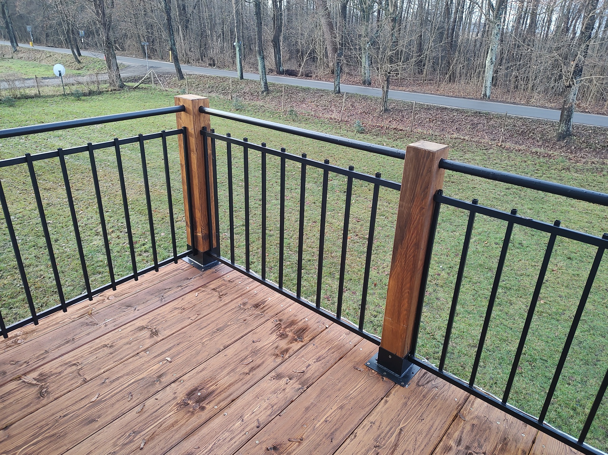Nowoczesny taras z desek w odcieniach brązu, zabezpieczony czarną, metalową balustradą z drewnianymi słupkami, widok na zielony trawnik i alejkę parkową w tle.