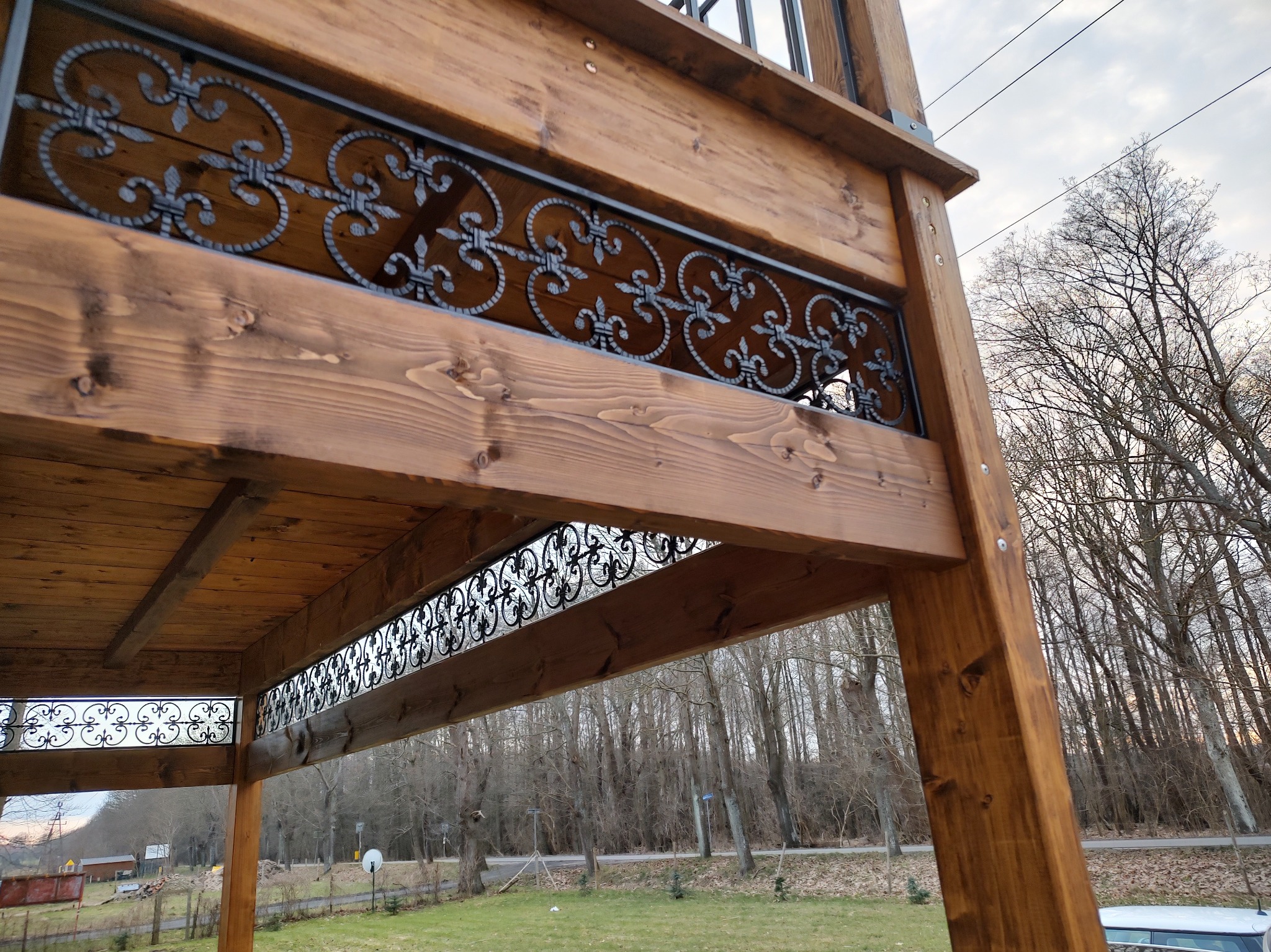 Drewniana konstrukcja tarasu z ozdobnymi, metalowymi balustradami, widok z dołu na tle drzew i pochmurnego nieba.