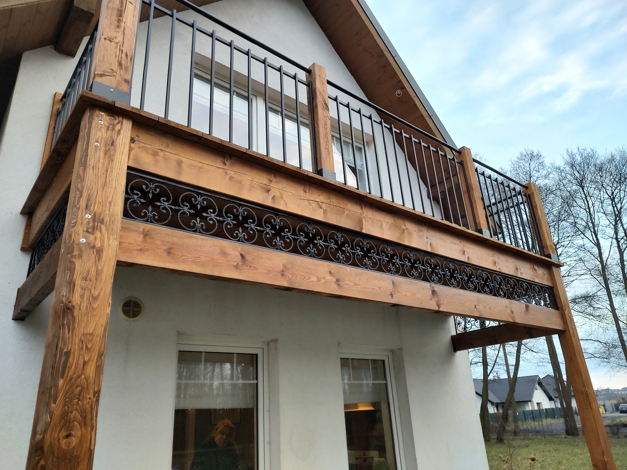 Drewniany balkon z czarną, kutą balustradą, wsparty na solidnych, impregnowanych belkach, widoczny z perspektywy dolnej, z fragmentem elewacji budynku w tle.