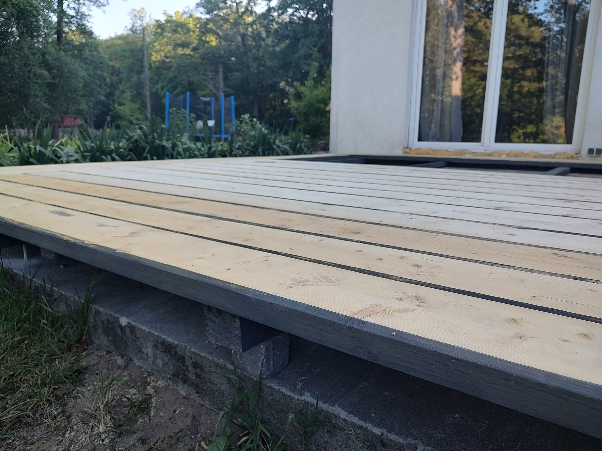 Montaż drewnianego tarasu: surowe deski tarasowe ułożone na ciemnoszarej konstrukcji, widoczny fragment elewacji budynku i trawnik z elementami placu zabaw w tle.