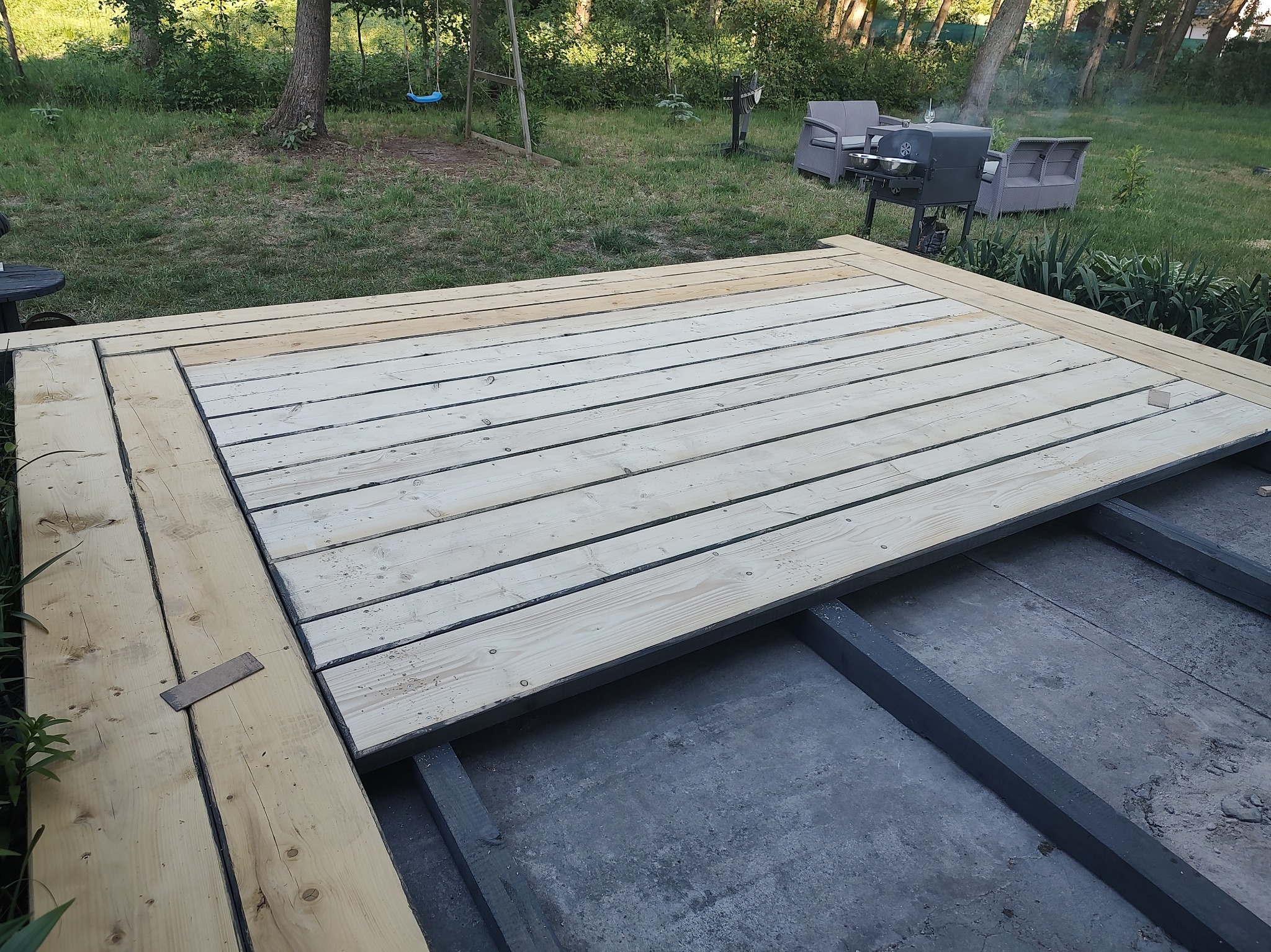 Budowa tarasu: deski ułożone na czarnej konstrukcji nośnej, w tle trawnik z grillem i meblami ogrodowymi.