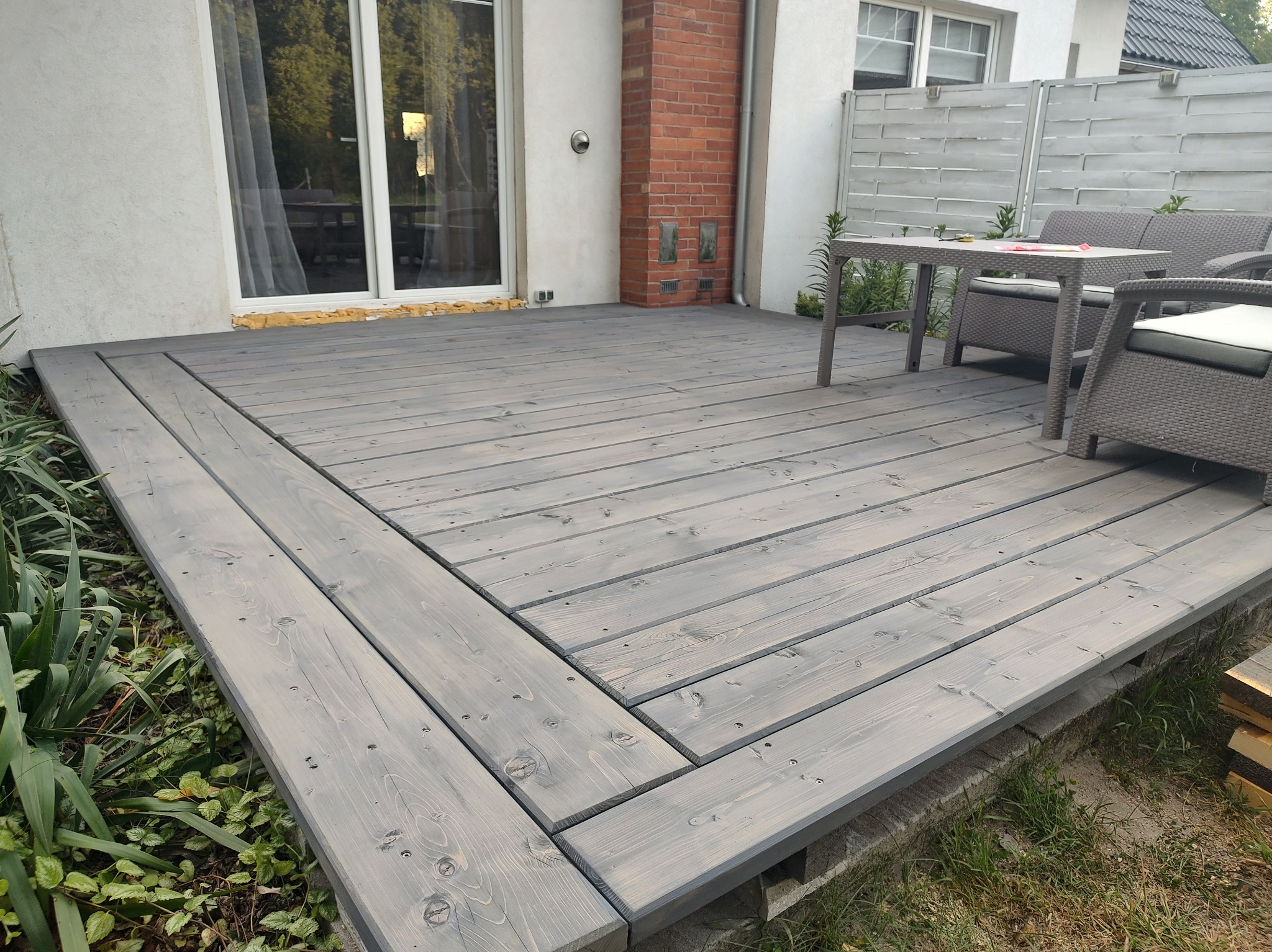 Szary taras z desek, z meblami ogrodowymi i widokiem na dom z oknami.