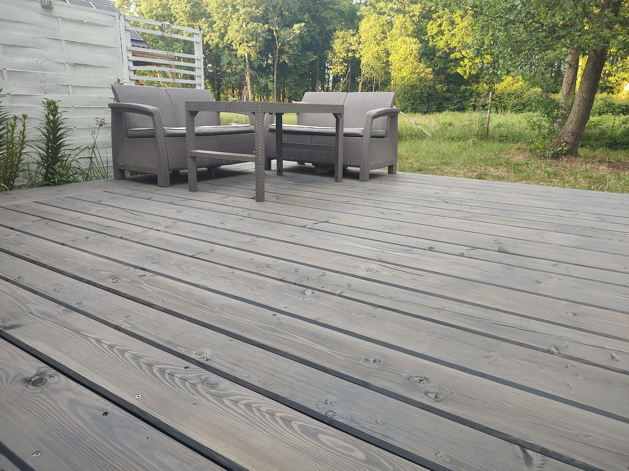 Szary taras z desek z widocznymi śrubami, na którym stoi zestaw mebli ogrodowych z technorattanu w odcieniach szarości, a w tle widać zieleń drzew i biały płot.