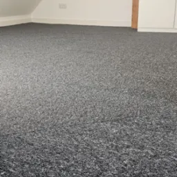 Szara wykładzina dywanowa w pokoju na poddaszu, widoczna ściana ze skosem, gniazdka elektryczne i drewniany element konstrukcyjny.