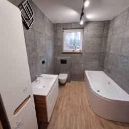 Marcin Tomaszewski Usługi Remontowe - Nowoczesna łazienka z szarymi płytkami, drewnopodobną podłogą, białą wanną narożną, umywalką z szafką w kolorze drewna i białym wysokim regałem, oświetlona reflektorami sufitowymi.