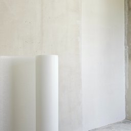 Marcin Tomaszewski Usługi Remontowe - Przygotowanie do malowania wnętrza: wiadro z farbą, wałek i rolka folii malarskiej w surowym pokoju z częściowo pomalowaną ścianą.