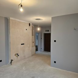 Marcin Tomaszewski Usługi Remontowe - Wnętrze mieszkania w trakcie remontu: ściany częściowo pokryte szpachlą, częściowo pomalowane na szaro, widoczne instalacje elektryczne i wodne, surowy beton na podłodze, wiszące żarówki.