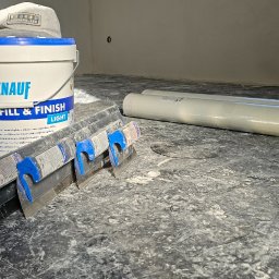 Marcin Tomaszewski Usługi Remontowe - Przygotowanie do szpachlowania ścian: wiadro z masą szpachlową Knauf Fill & Finish Light, szpachelki z niebieskimi uchwytami, zakryty folią podkład, oraz czapka leżąca na wiadrze, na podłodze...