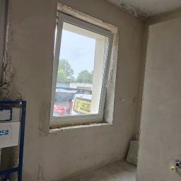 Marcin Tomaszewski Usługi Remontowe - Surowa łazienka w trakcie remontu: ściany w szarym tynku, widoczne okna z białymi ramami, zamontowany stelaż podtynkowy WC marki Roca.