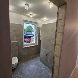 Marcin Tomaszewski Usługi Remontowe - Łazienka w trakcie remontu z szarymi płytkami imitującymi marmur, widoczne surowe wykończenie framugi drzwi, biała toaleta i okno z widokiem na podwórko z czerwonym samochodem.