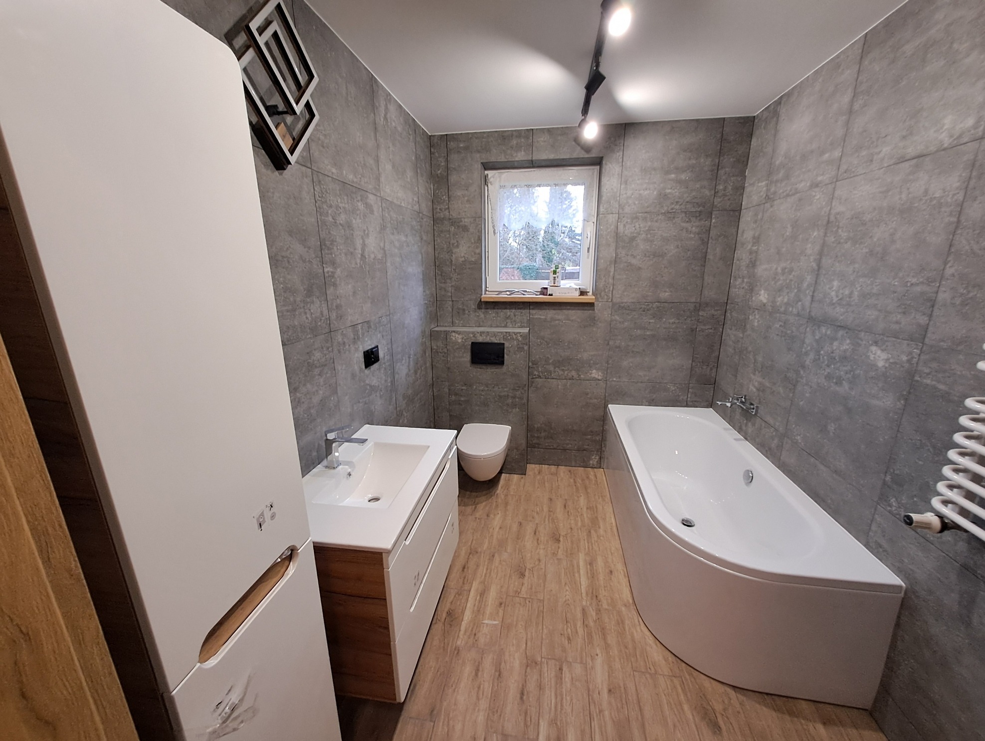 Nowoczesna łazienka z szarymi płytkami, drewnopodobną podłogą, białą wanną narożną, umywalką z szafką w kolorze drewna i białym wysokim regałem, oświetlona reflektorami sufitowymi.