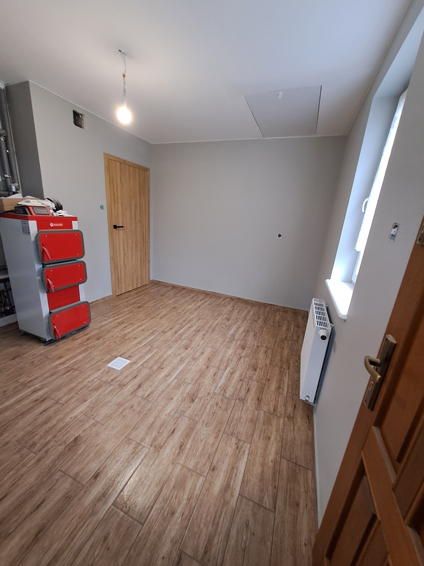 Wnętrze pokoju z drewnopodobnymi panelami podłogowymi, czerwono-szarym piecem CO z widocznymi rurami, grzejnikiem pod oknem i drewnianymi drzwiami z czarną klamką.