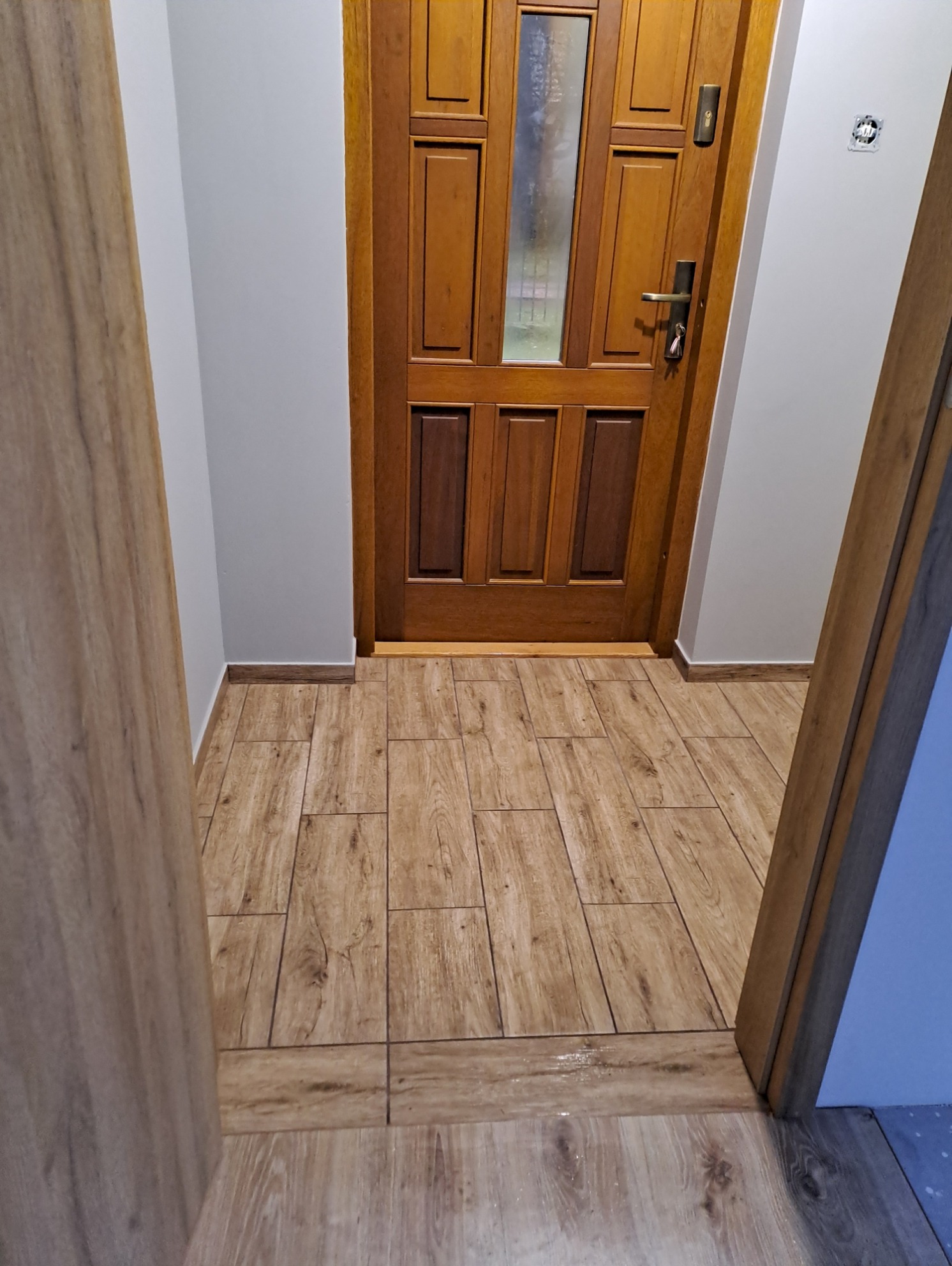 Widok na drewnianą podłogę z płytek imitujących drewno, dopasowaną do drewnianych drzwi wejściowych z szybą i futryną, w korytarzu o szarych ścianach.