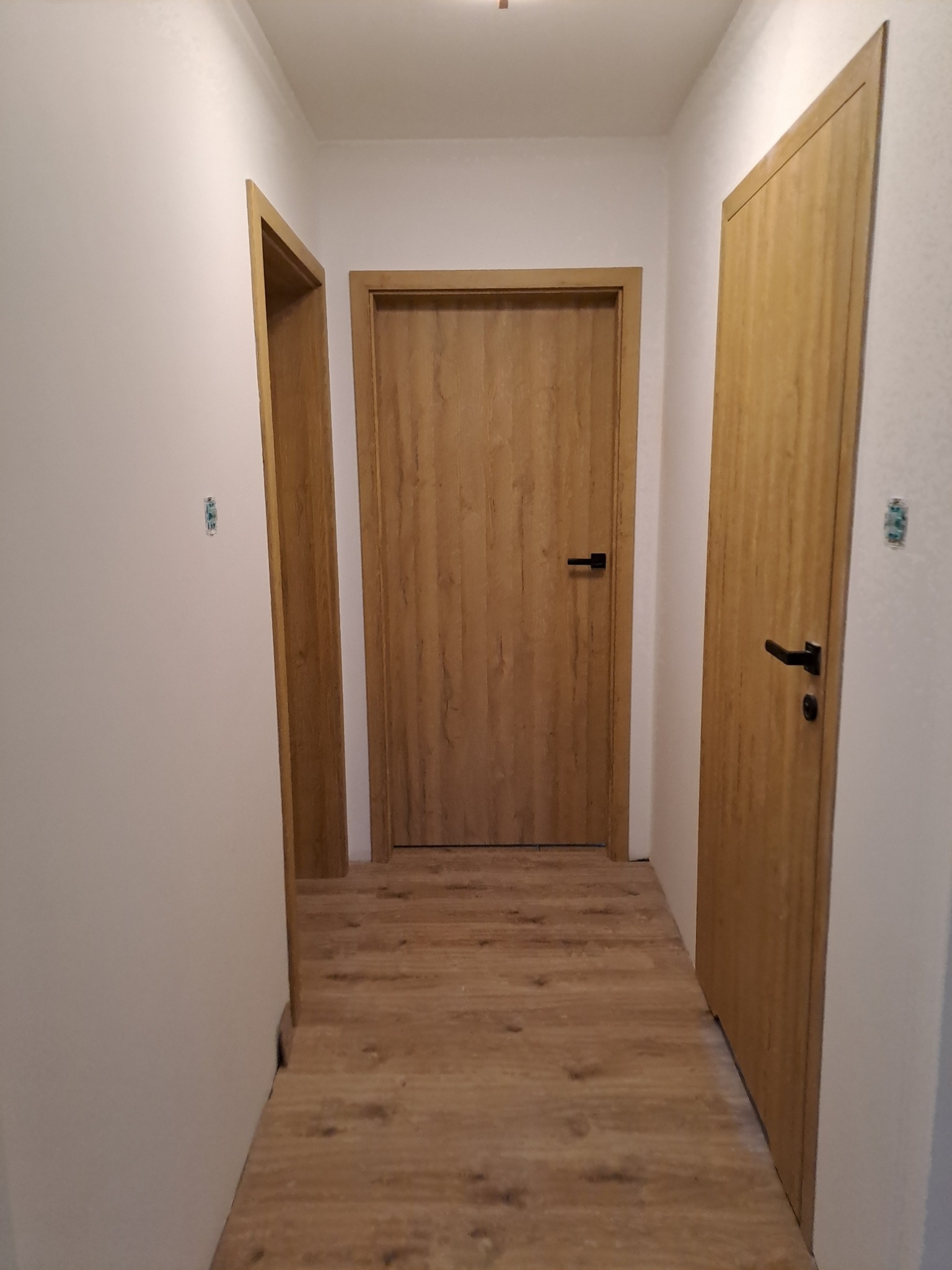 Korytarz po remoncie z trzema drewnianymi drzwiami o różnych szerokościach, czarnymi klamkami i panelami podłogowymi w odcieniach brązu.