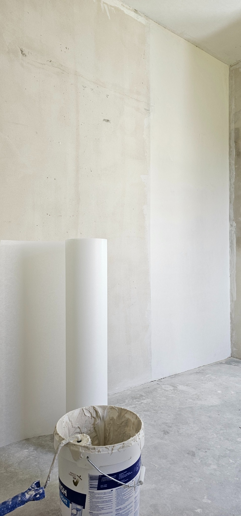 Przygotowanie do malowania wnętrza: wiadro z farbą, wałek i rolka folii malarskiej w surowym pokoju z częściowo pomalowaną ścianą.