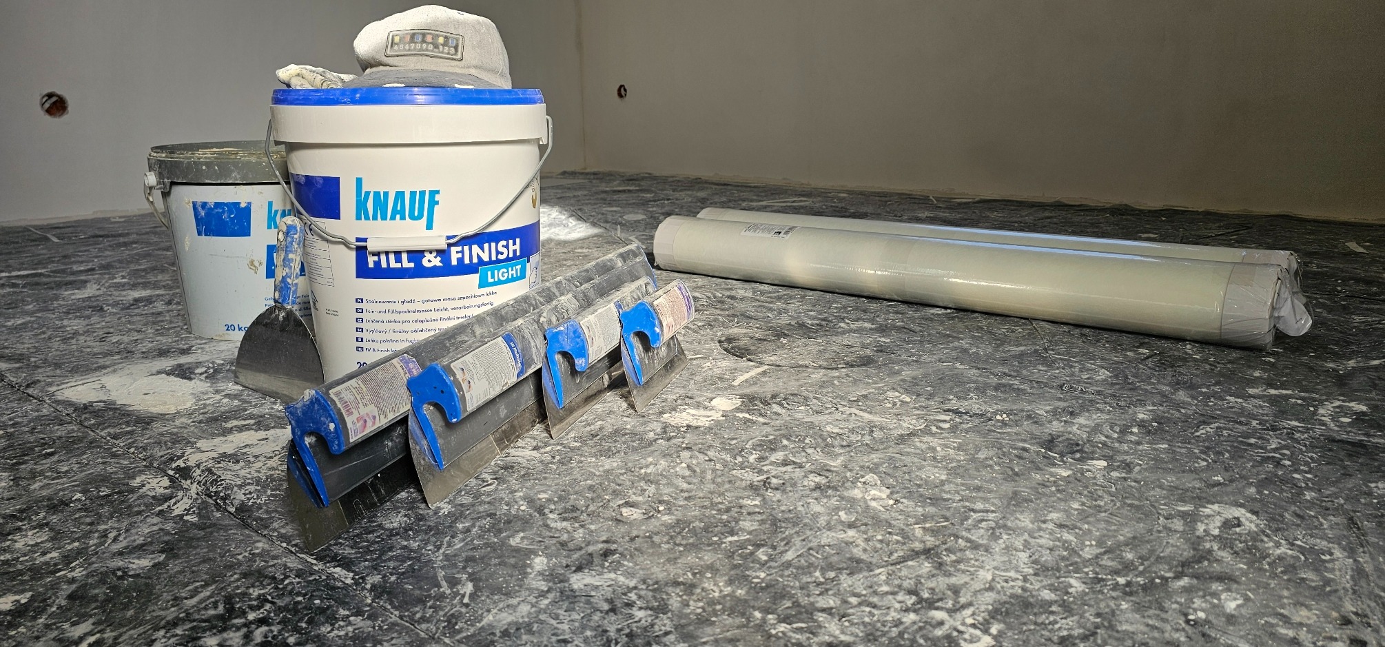 Przygotowanie do szpachlowania ścian: wiadro z masą szpachlową Knauf Fill & Finish Light, szpachelki z niebieskimi uchwytami, zakryty folią podkład, oraz czapka leżąca na wiadrze, na podłodze...
