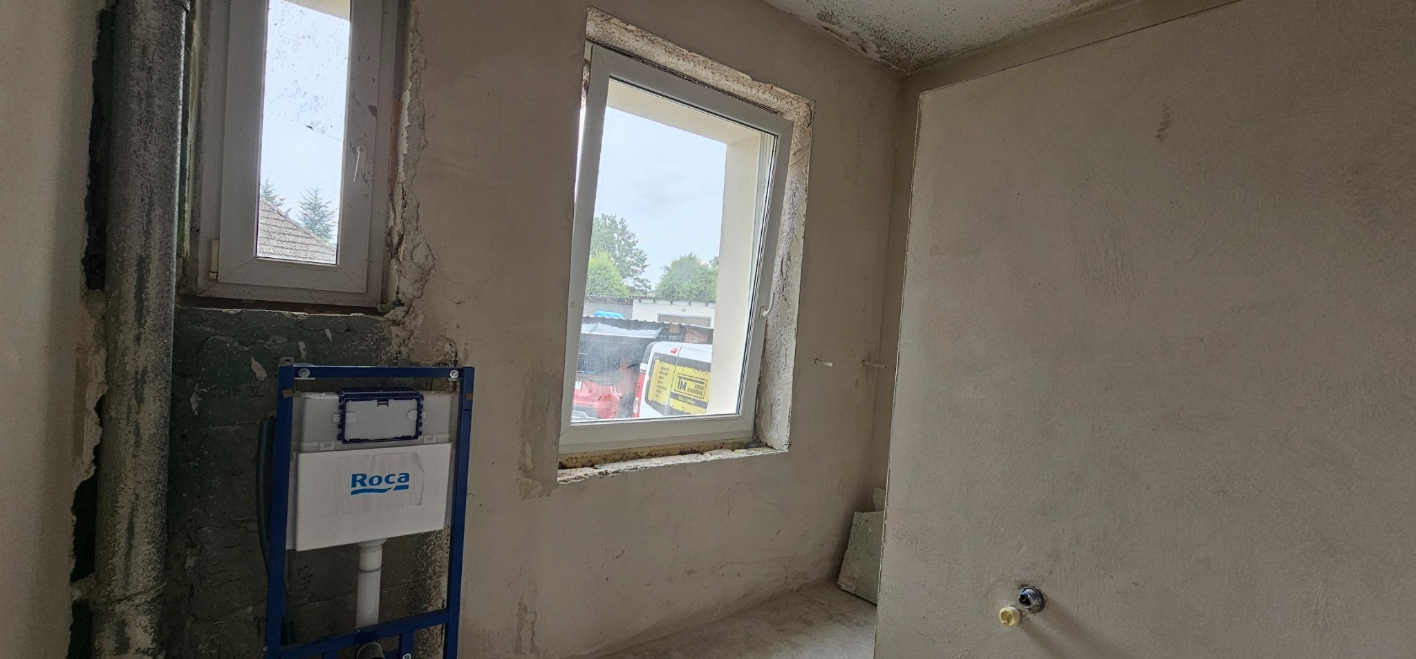Surowa łazienka w trakcie remontu: ściany w szarym tynku, widoczne okna z białymi ramami, zamontowany stelaż podtynkowy WC marki Roca.
