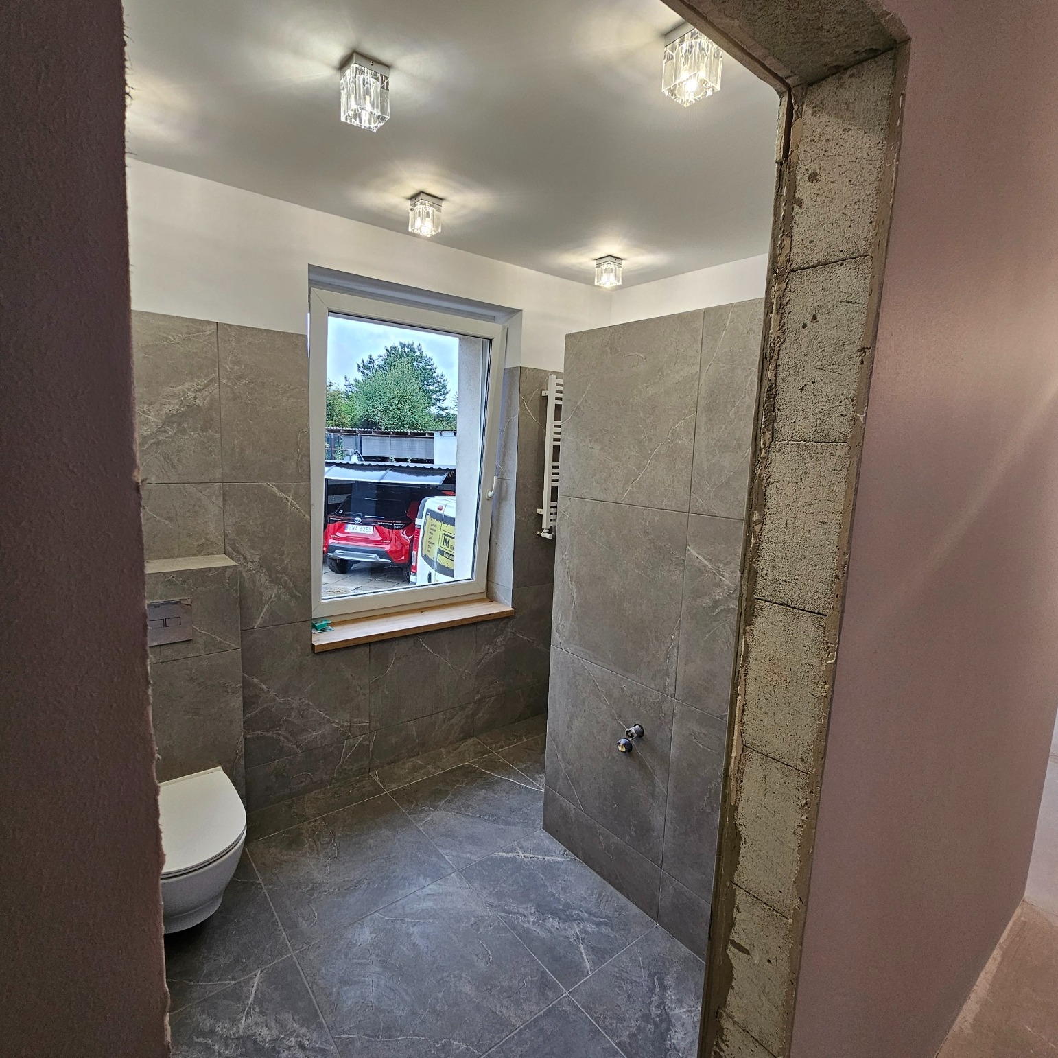 Łazienka w trakcie remontu z szarymi płytkami imitującymi marmur, widoczne surowe wykończenie framugi drzwi, biała toaleta i okno z widokiem na podwórko z czerwonym samochodem.
