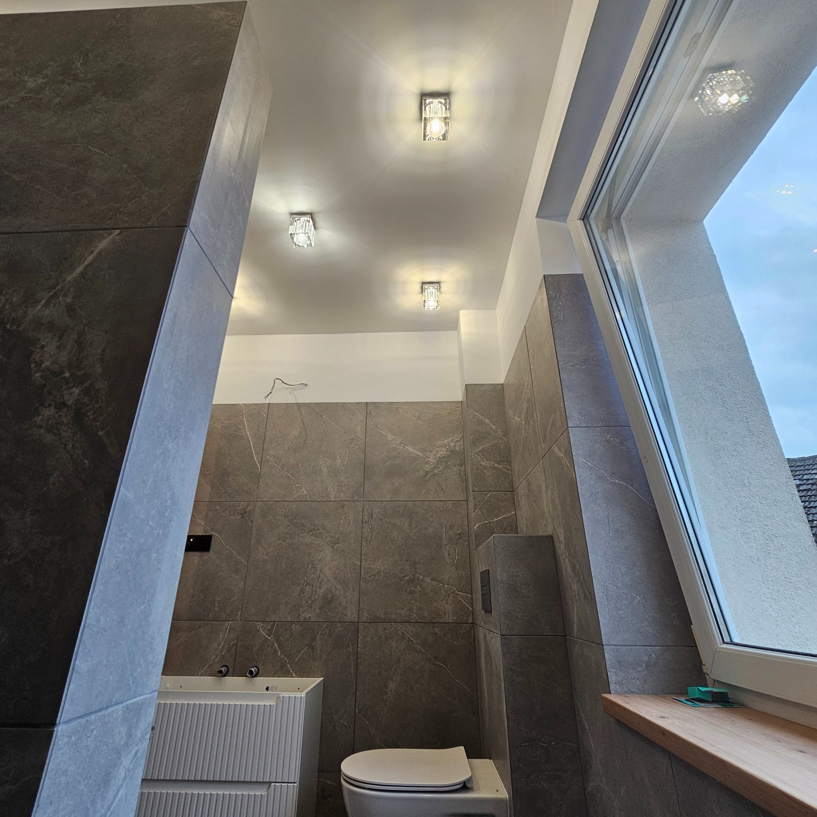 Nowoczesna łazienka w szarościach z marmurowymi płytkami, trzema kryształowymi lampami sufitowymi i oknem dachowym z drewnianym parapetem, częściowo widoczna biała szafka umywalkowa i toaleta.