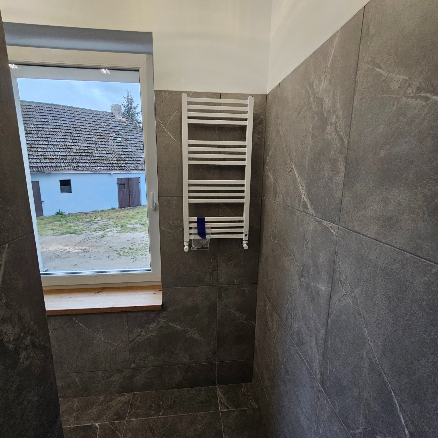 Wykończona łazienka z szarymi płytkami imitującymi marmur, białym grzejnikiem drabinkowym i widokiem na wiejski dom przez okno.