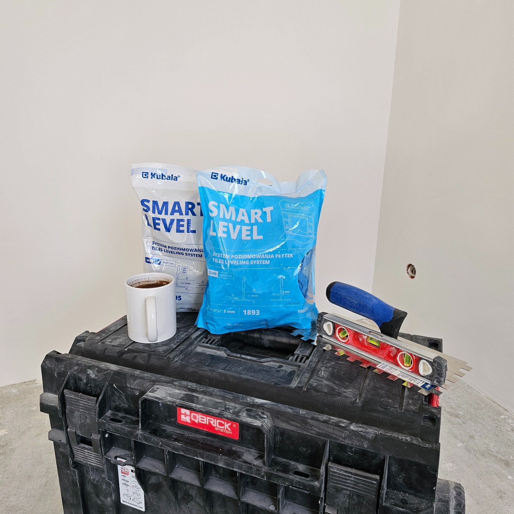 Dwa opakowania systemu poziomowania płytek Kubala Smart Level, poziomica i kielnia ząbkowana leżące na czarnym kufrze narzędziowym Qbrick, obok biały kubek z kawą, w tle surowa ściana przygotowana...