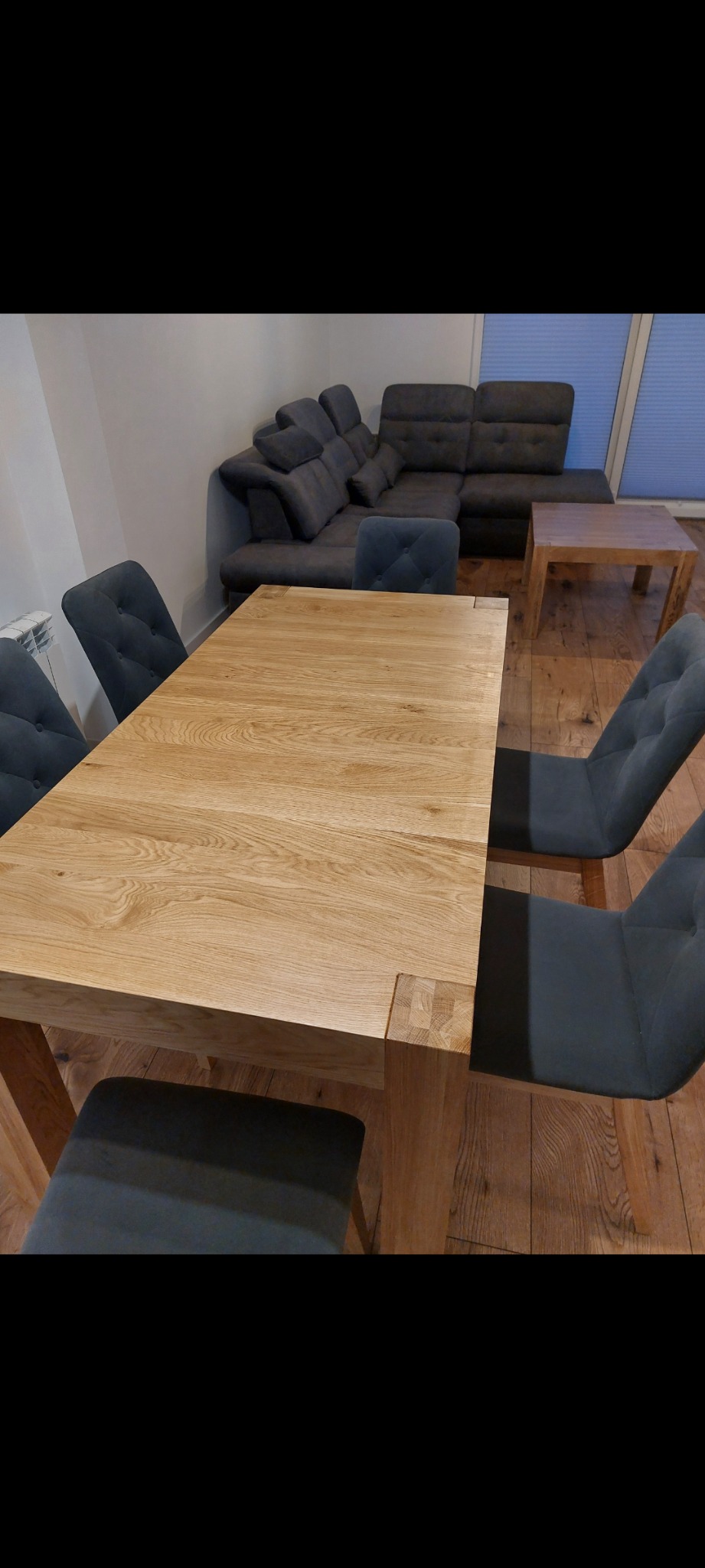 Jasny, rozkładany stół z litego drewna z czterema tapicerowanymi krzesłami w ciemnym odcieniu szarości, ustawiony w salonie z parkietem i szarą sofą narożną w tle.