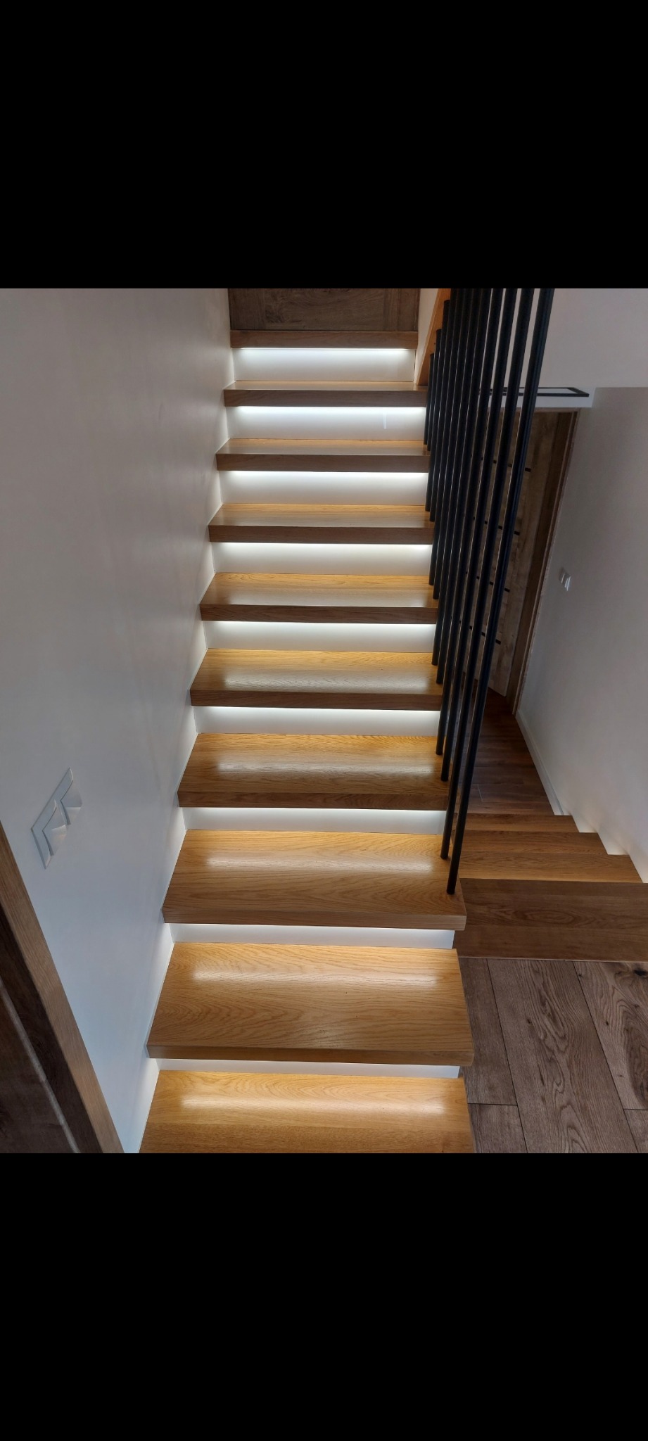 Nowoczesne schody drewniane z podświetleniem LED pod każdym stopniem i czarną, minimalistyczną balustradą, widok z dołu w górę, z drewnianą podłogą w tle.