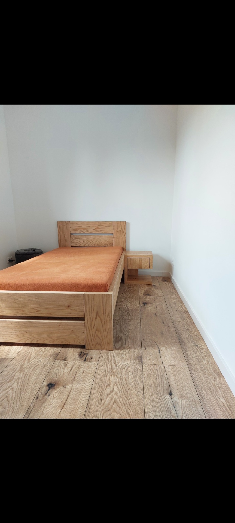 Nowoczesne drewniane łóżko z pomarańczowym materacem i pasującą szafką nocną, ustawione na podłodze z desek o naturalnym odcieniu.