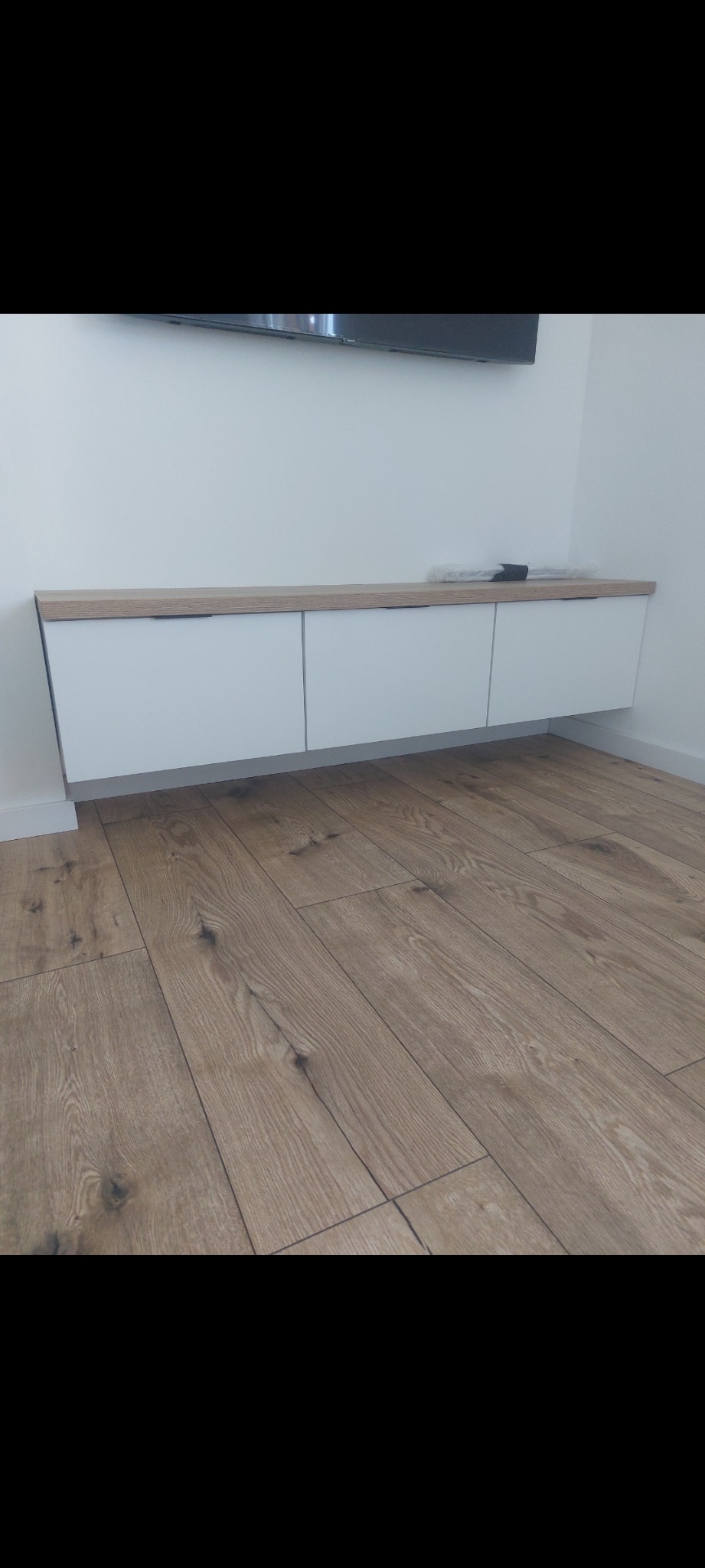 Minimalistyczna szafka RTV wisząca na ścianie, z białymi frontami i blatem w kolorze jasnego drewna, zamontowana nad podłogą z paneli w odcieniach brązu.