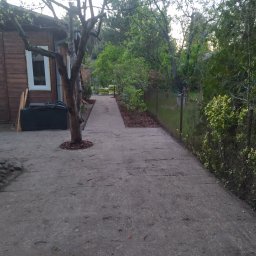 Usługi Ogrodnicze Marcin Oliński - Betonowa ścieżka prowadząca przez ogród w kierunku drewnianego domku, z drzewem posadzonym w wyciętym otworze w kostce i podsypanym korą.